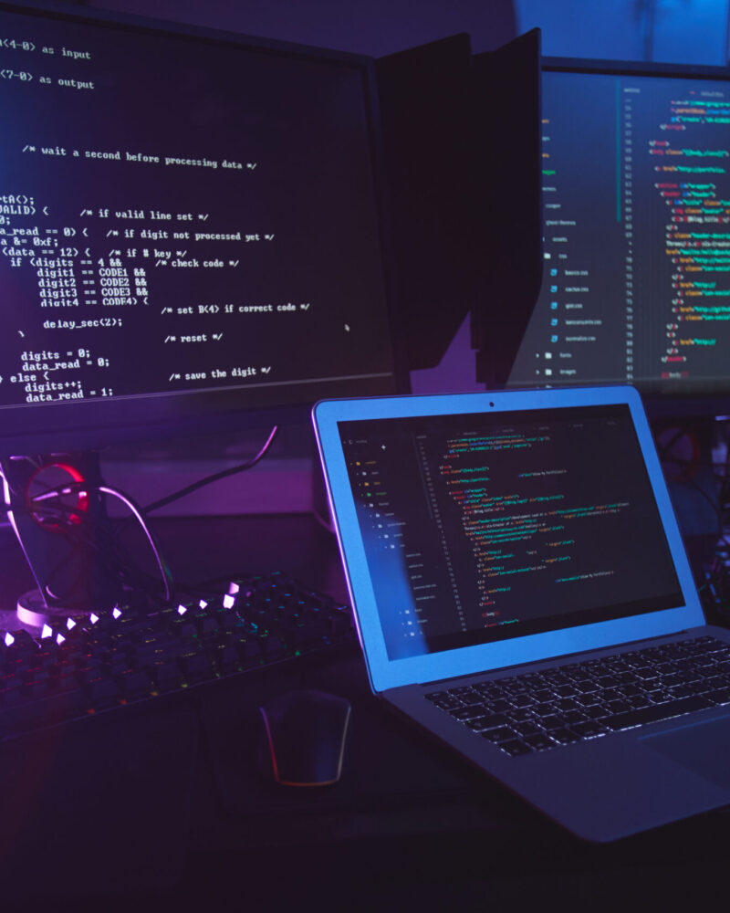 Background image of various computer equipment with programming code on screens on table in dark room, cyber security concept, copy space